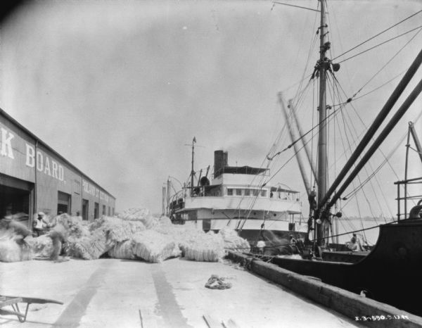 View down dock towards bales of manila being unloaded from a ship. The building on the left has a sign that reads: "Poland Street Wharf." There is a ship at dock on the right.