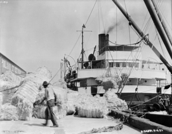 Bales of manila are lying on a dock after being unloaded from a ship, which is at dock on the right. The building on the left has a sign that reads: "Poland Street Wharf." 