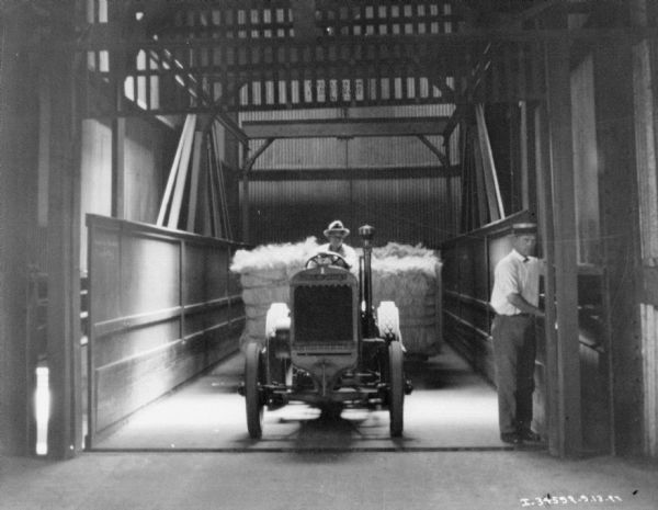 View towards man sitting behind the driver's wheel of a McCormick-Deering tractor in what may be an elevator. Behind the tractor is a cart filled with bales of manila. A man is standing and operating the elevator on the right.