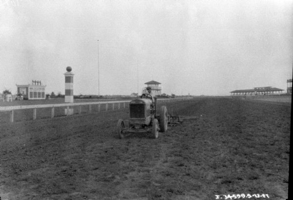 View down race track towards a man driving a tractor pulling disk harrows to smooth the track. In the background are buildings and viewing stands.