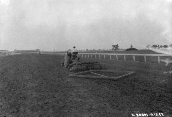 Rear view of a man using a tractor to pull a disk harrow to smooth out a racetrack.