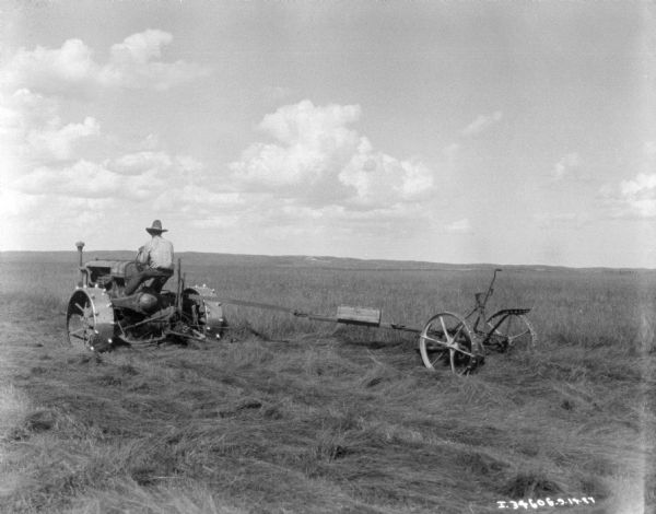 Three-quarter view from rear left of a man using a Farmall tractor to pull a mower with a modified hitch.