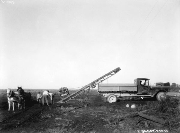 View across field towards men working an irrigation operation. On the right is a team of horses. The men are working nearby near a piece of machinery set up behind a truck. A man is sitting in the driver's seat of the truck, and painted on the passenger door of the truck is a sign that reads: "Lucas Bros."
