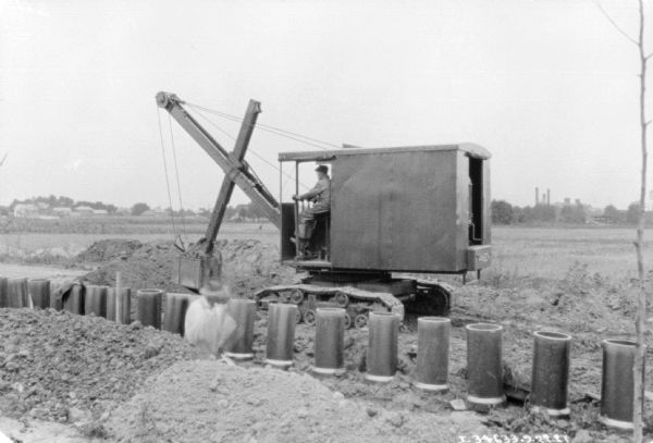In the foreground a man is working near a row of pipes. Behind him a man is operating an earthmover.