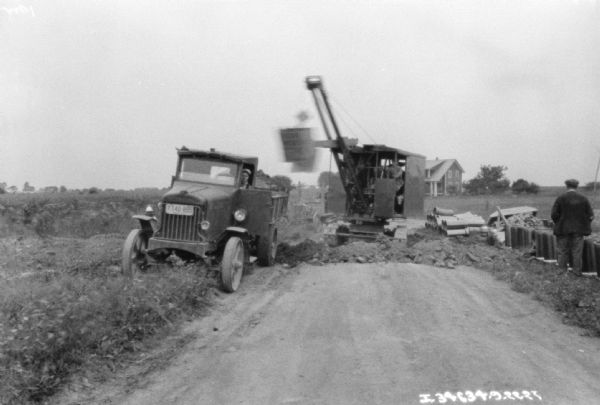 View down road towards a man using an earthmover for road repair. There is a truck parked on the roadside on the left, and a stack of pipes is near the road on the right.
