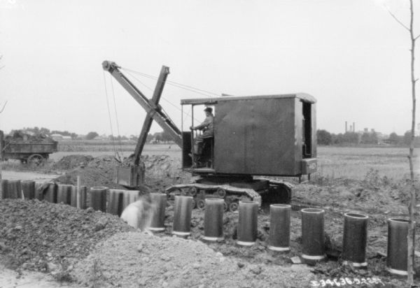 In the foreground a man is working near a row of pipes. Behind him a man is operating an earthmover.