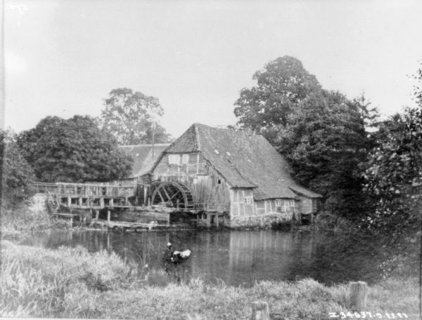 View from grassy shoreline towards a watermill with a thatched roof.