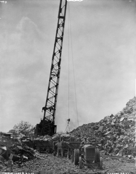A man is sitting on an industrial tractor that is pulling an open wagon. More wagons are behind, and men are directing a crane to either load or unload the wagons. Large piles of material are on the left and right.