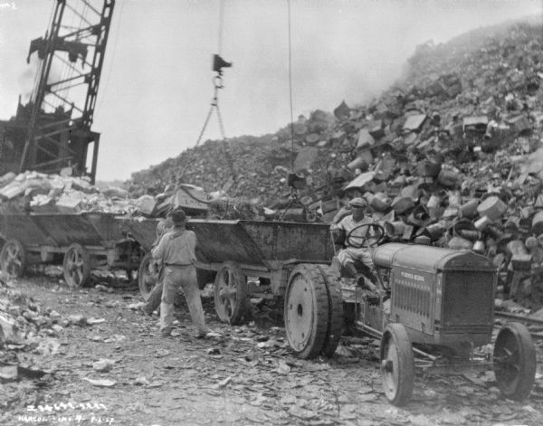 A man is sitting on an industrial tractor that is pulling an open wagon. More wagons are behind, and men are directing a crane to either load or unload the wagons. Large piles of material are on the left and right.