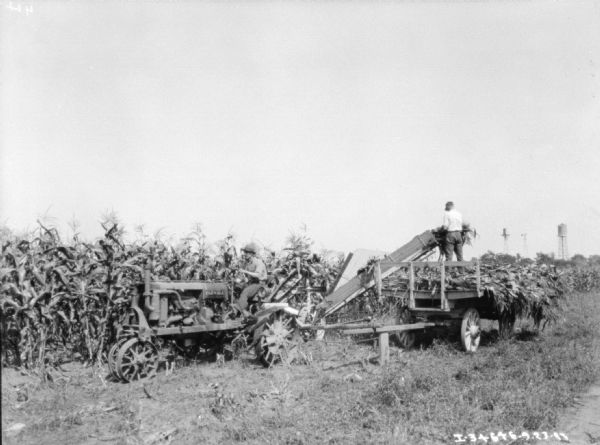 View towards a man driving a Farmall tractor pulling a corn picker in a cornfield. Another man is standing on a wagon pulling cornstalks out of the picker. The wagon is piled with cornstalks. There are water towers in the background on the right.