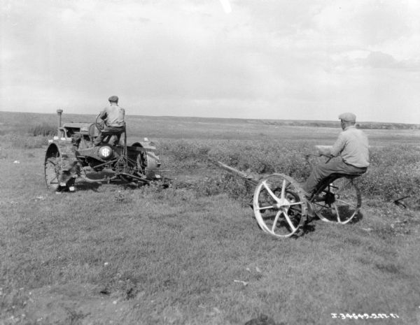 Three-quarter view from left rear of a man driving a Farmall tractor pulling another man on a mower in a field.