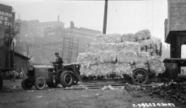 Men are unloading bales for twine stacked on a wagon. Industrial buildings are in the background.