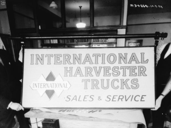 A man (face out of frame) on the left is holding up an International Harvester Trucks Sales & Service sign resting on a table. Another person (face out of frame) is holding the sign on the right.