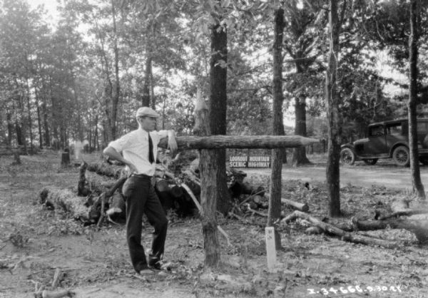 A man is standing at a signpost made of logs in the shape of an arrow. A sign along a road in the background reads: "Lookout Mountain Scenic Highway." There is an automobile parked near the sign on the right.