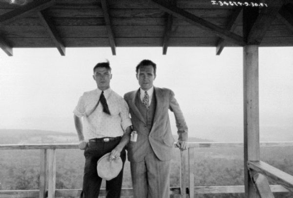 Two men are posing against a wood railing. Over their heads is a roof. Behind them is a view of wooded hills.