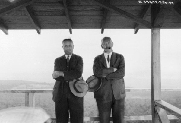 Two men are posing against a wood railing. Over their heads is a roof. Behind them is a view of wooded hills.