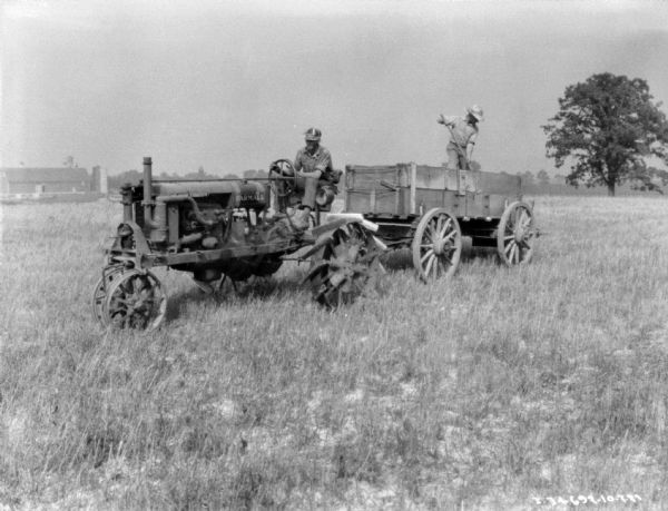 Three-quarter view from front left of a man driving a Farmall tractor pulling a lime spreader in a field. A man is standing in the lime spreader holding a shovel. There are farm buildings in the background.