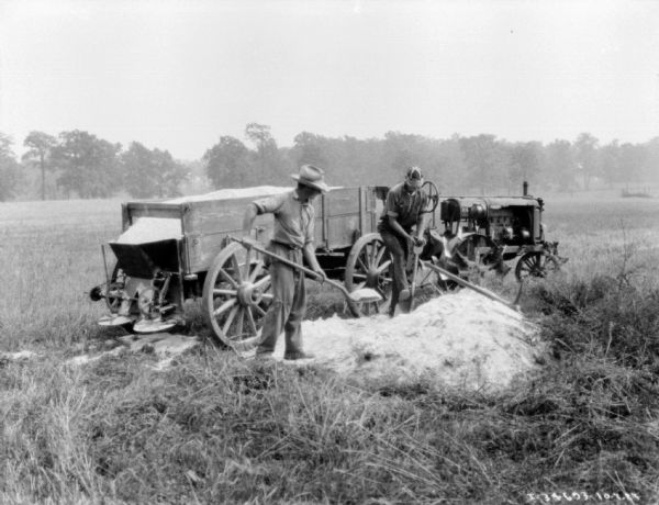 Two men are holding with shovels to spread a pile of lime in a field. Behind them is a Farmall tractor pulling a lime spreader.