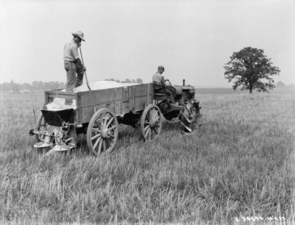 Three-quarter view from right rear of a man driving a Farmall tractor pulling a lime spreader in a field. A man is standing in the lime spreader holding a shovel. 