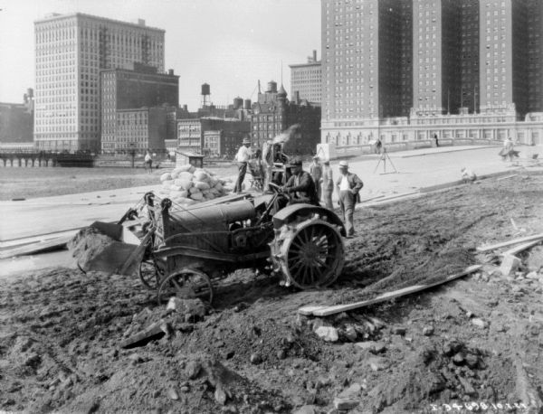 A man is operating an industrial tractor, which has a shovel on the front for moving earth. Men are in the background standing near a pile of sandbags. Tall buildings, a bridge, and water towers are in the background.
