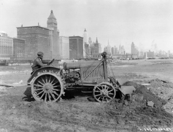 A man is operating an industrial tractor, which has a shovel on the front for moving earth. Tall buildings are in the background.