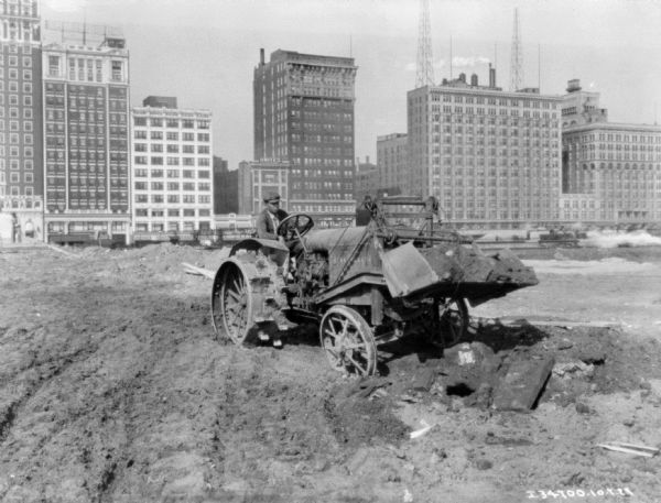 A man is using an industrial tractor, which has a shovel on the front for moving earth. Tall buildings are in the background.