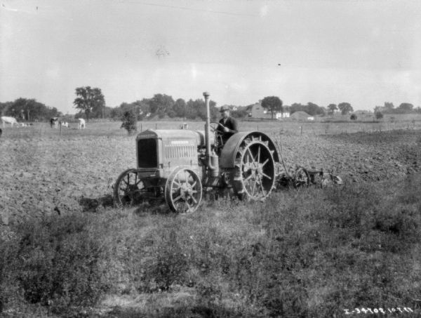 Three-quarter view from front left of a man driving a tractor pulling a plow in a field. In the background are cows in a field, and farm buildingss are in the far distance.