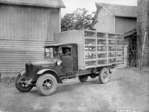 A man is sitting in the driver's seat of a delivery truck for chickens. Barns and a silo are in the background.
