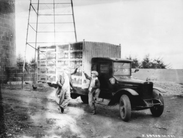 Two men are standing next to the side of a livestock delivery truck for chickens. There is a silo in the background, and the metal base of what may be a water tower.