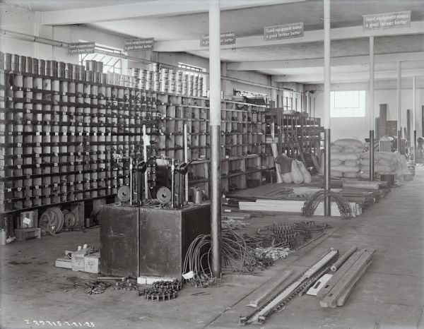 Repair shop at a dealership, with tools, parts and workstations. On the left are tall shelves with compartments and cubbies for storing parts, with stacks of cans of oil and grease along the top. Light is coming through windows in the walls in the background. Signs hanging from the ceiling read: "Good equipment makes a good farmer better."
