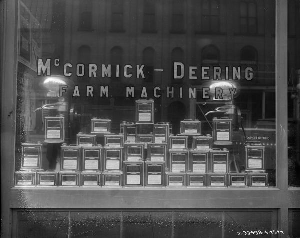 Exterior view of a display window of cans of McCormick-Deering Cream Separator Oil, with cream separators in the background at a dealership. A sign painted on the window reads: "McCormick-Deering, Farm Machinery." Reflected in the window are brick buildings directly across the street.