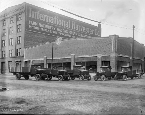 View across cobblestone street towards men sitting in the cabs of six International trucks lined up in two rows. Behind them is a two-story brick building with the IHC logo at the roof line, and trucks on display behind large show windows along the sidewalk in front. The five-story brick building next door on the left has a sign painted on the side that reads: "International Harvester of America."