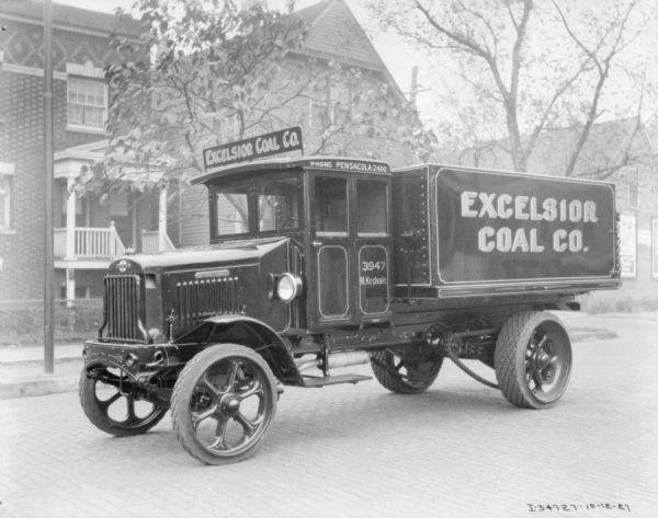 View across cobblestone street towards a coal delivery truck parked in front of buildings. The sign above the cab reads: "Excelsior Coal Co."