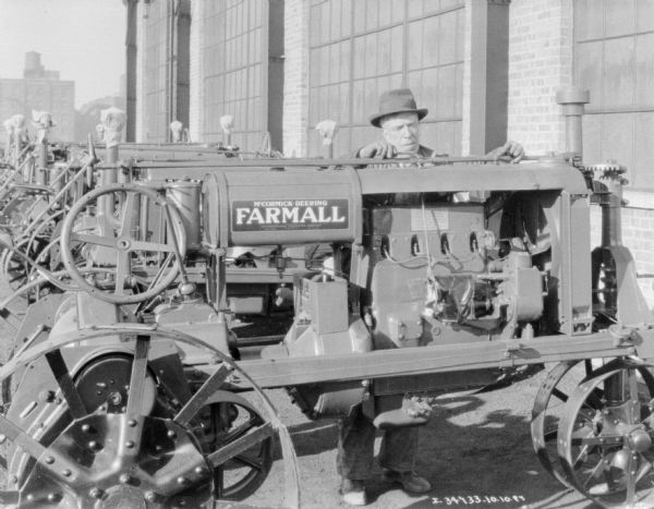 A man is standing outdoors behind a Farmall tractor making adjustments with a wrench. Behind him is a brick building.