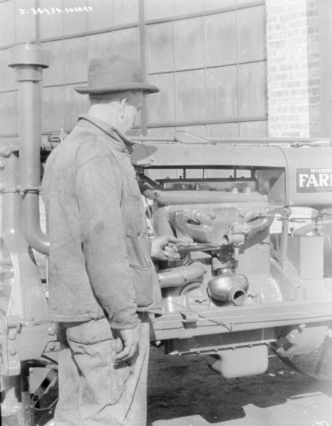 A man is holding a tool against a Farmall tractor engine while standing outdoors in front of a brick building at a dealership.