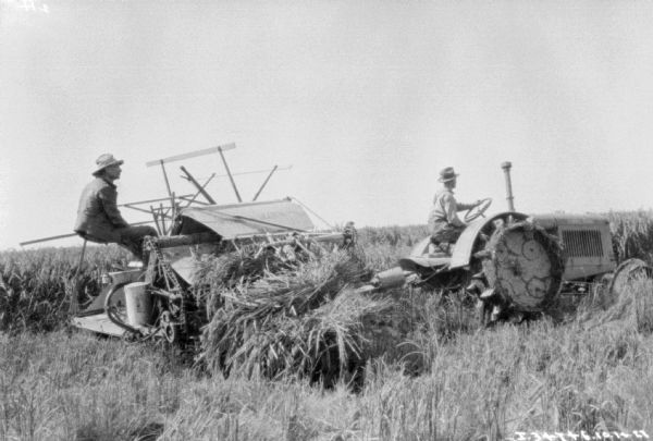 View of a man driving a McCormick-Deering tractor pulling a man on a binder in a field.