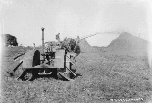 View towards the rear of a tractor in the foreground. In the background two men are working to load a harvester-thresher which is shooting grain onto a large pile in a field. Another man is standing and watching them on the left behind the tractor.