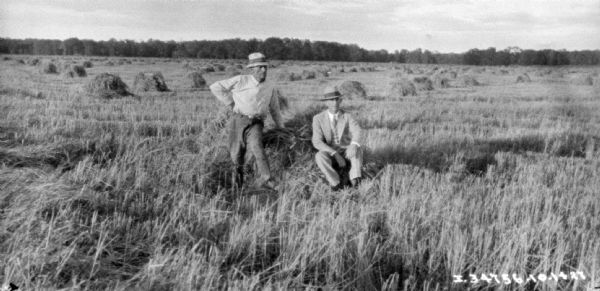 Two men are in a field with shocks of grain dotting the field. One man is wearing a suit and hat. Trees are in the background.