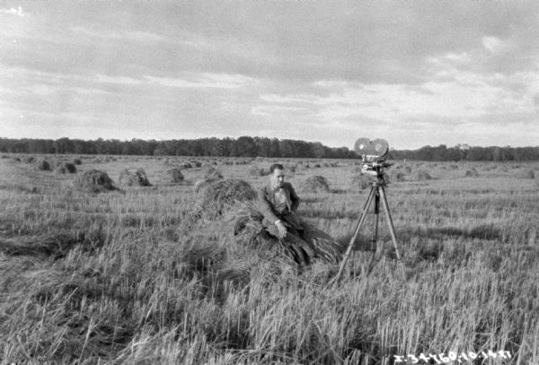 A young man is sitting on a stack of hay or grain in a field near a movie camera on a tripod.