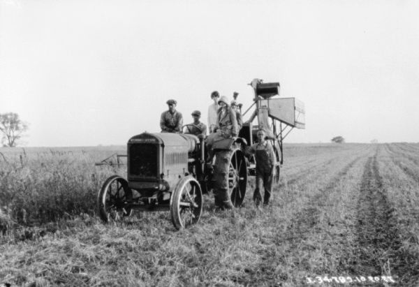 View of a group of men and women posing on a McCormick-Deering tractor pulling a harvester-thresher.