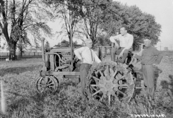 Three men are posing with a Farmall tractor in a field. In the background are trees, fence, road, and farm buildings. There is a truck behind the group on the right.