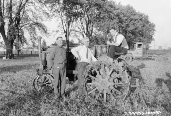 Three men are posing with a Farmall tractor in a field. In the background are trees, fence, road, and farm buildings. There is a truck behind the group on the right.