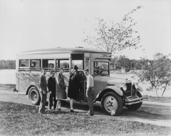 View across lawn towards a group of teenagers boarding a small bus parked on a road with a body of water in the background.