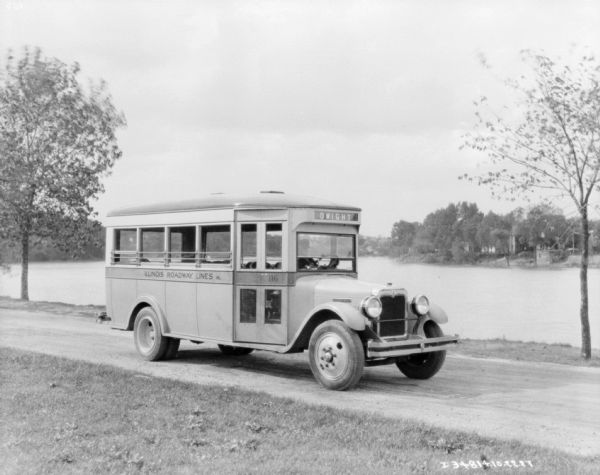 View across lawn towards a small bus parked on a road with a body of water in the background. There is a man sitting in the driver's seat.