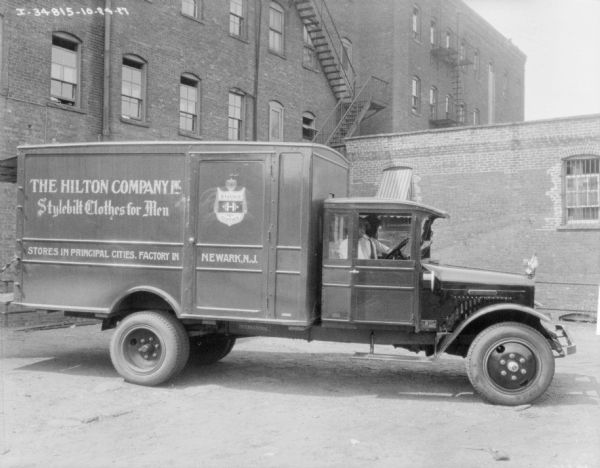 Right side profile view of two men sitting in the cab of a delivery truck parked in front of brick buildings. The sign painted on the side of the truck reads: "The Hilton Company, Inc., Stylebilt Clothes for Men."