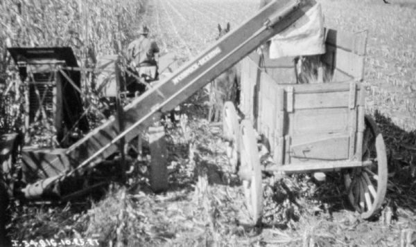 Elevated rear view of a man driving a tractor pulling a McCormick-Deering corn picker in a field. On the right is a horse-drawn wagon.