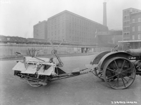 A tractor is hitched to a McCormick-Deering binder parked on the brick pavement. Railroad cars are on elevated railroad tracks in front of factory buildings in the background.