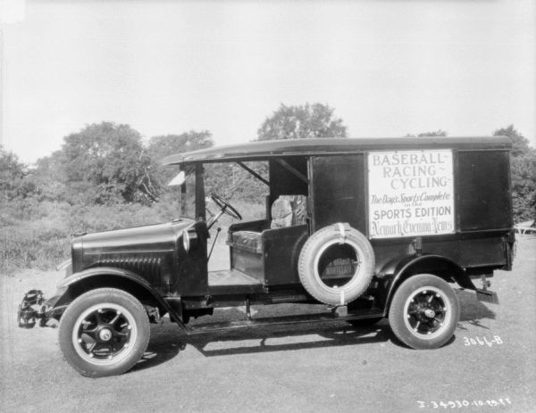Left side view of a delivery truck parked outdoors on a road. The cab has no doors, and the front seat is one long bench with a cushion. The sign on the back of the enclosed truck bed reads: "Baseball — Racing — Cycling, The Day's Sports Complete in the Sports Edition Newark Evening News."