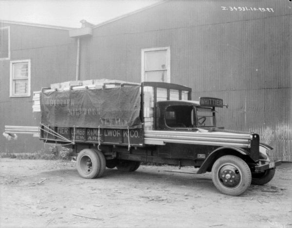 A delivery truck parked near the side of the building. A sign above the windshield of the truck reads: "Whittier." Lumber is stacked in the bed of the truck, and long pieces of lumber extend out from the back of the truck alongside the passenger door to the front of the truck. A tarp on the side of the truck is painted with a sign that reads: "Whittier Millwork Newark."