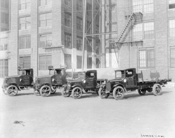 Four delivery trucks are parked in a line at an angle. Behind the trucks are industrial brick buildings with fire escapes.
